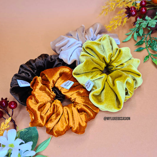 Four velvet scrunchies in brown, orange, beige, and mustard yellow on a peach background with floral decorations.