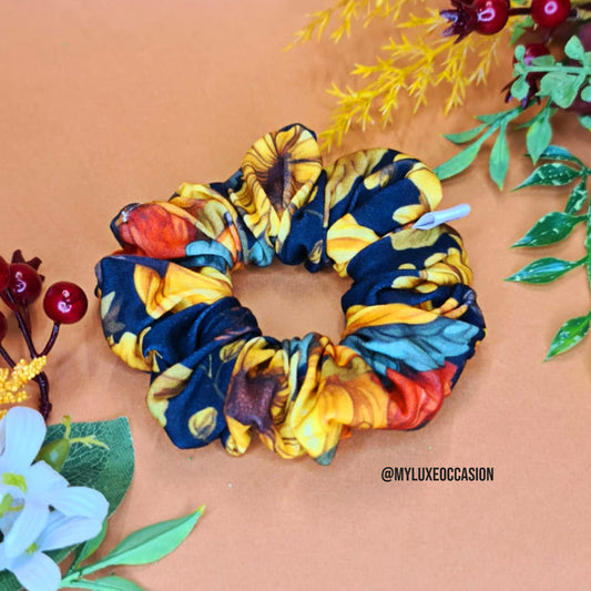 Black pumpkin floral scrunchie on a beige background with decorative plants.